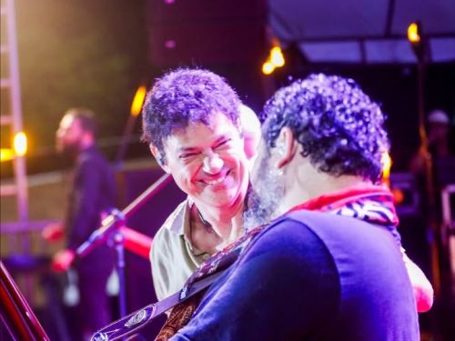 Foto no palco mostrando Jorge Vercillo sorrindo para outro músico, que está de costas tocando guitarra. O ambiente é de show, com luzes de fundo e equipamentos de som visíveis.