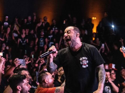 Fotografia em plano médio focada na expressão do cantor. Badauí está de frente, com os olhos fechados e a boca aberta em um grito potente enquanto segura o microfone. Sua camiseta preta tem um escudo do Corinthians com a frase "Só para loucos". No canto inferior, fãs próximos à grade aparecem com expressões de euforia e braços levantados.