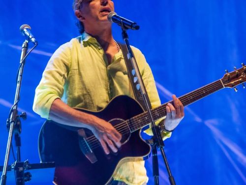 Foto colorida do cantor Zeca Baleiro tocando violão e cantando no palco. Ele veste uma camisa verde clara, calça jeans e boné, sob uma forte iluminação azul ao fundo.