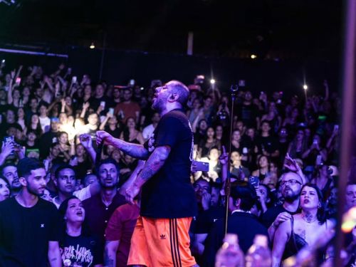 A fotografia registra um show da banda CPM 22 sob a perspectiva do palco para a plateia. Em destaque, o vocalista Badauí aparece de perfil, cantando com intensidade. Ele veste uma camiseta preta, bermuda esportiva laranja e exibe os braços tatuados.