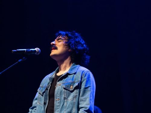 Fotografia em close de André Abreu durante uma performance. Ele está de perfil, com os olhos fechados e uma expressão de entrega emocional, posicionado diante de um microfone. Veste uma jaqueta jeans sobre camiseta preta, sob uma iluminação cênica azul que destaca seu rosto e cabelo ondulado.