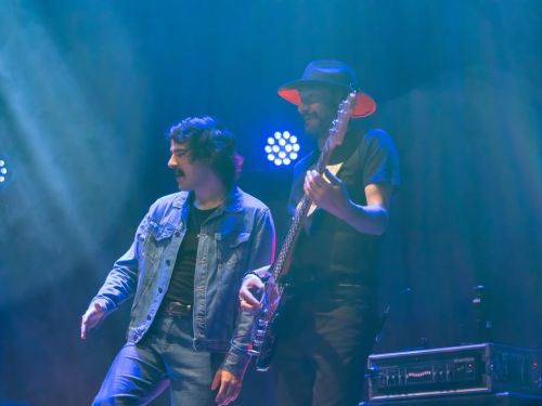 Fotografia de um show ao vivo em um palco iluminado por luzes azuis. À esquerda, André Abreu veste jaqueta e calça jeans, gesticulando enquanto canta. Ao seu lado, um músico de chapéu toca baixo elétrico. Ao fundo, luzes de LED e fumaça cênica completam o cenário artístico.