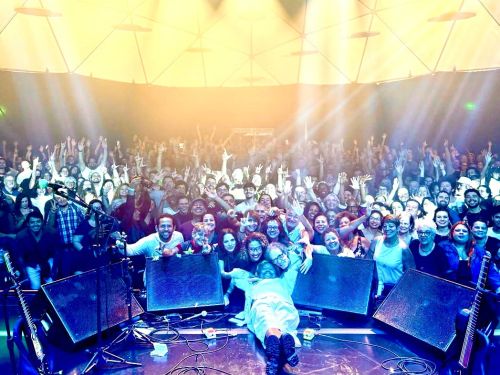 Foto ampla tirada do palco mostrando Chico César deitado à frente de uma plateia numerosa e animada que faz gestos com as mãos. A iluminação é forte e amarelada.