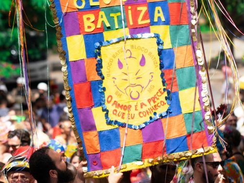 Foto mostra estandarte colorido do Bloco do Batiza, feito em patchwork, sendo erguido por foliões em meio a uma multidão animada no Parque do Palácio, durante um evento de carnaval ao ar livre.