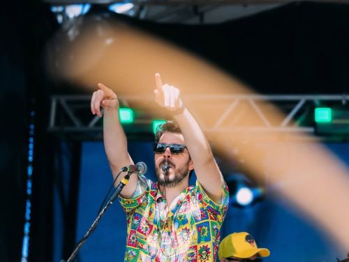 Homem de óculos escuros e camisa colorida canta no palco com os braços erguidos e dedos apontados para cima. Ao fundo, luzes de palco verdes e um baixista de boné amarelo. Uma faixa borrada de luz atravessa a frente da imagem.