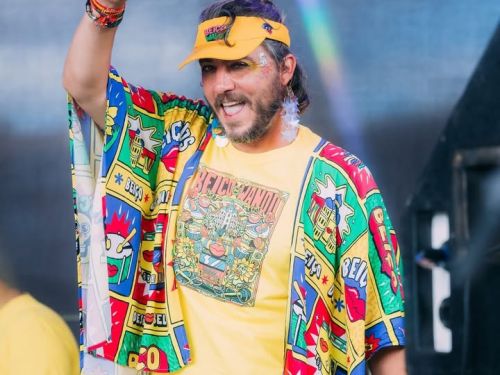 Homem sorridente com barba e viseira amarela aponta para cima. Ele veste um conjunto amarelo estampado com ícones coloridos do bloco "Beiço do Wando" e brincos de estrelas. O fundo é neutro e desfocado.