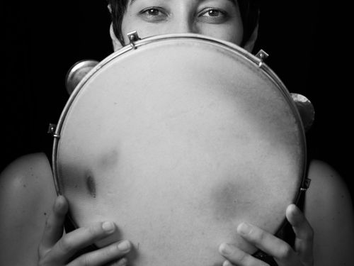 Fotografia artística em preto e branco de uma mulher (Analu Braga) com cabelos curtos e escuros. Ela segura um pandeiro circular à frente do rosto, cobrindo do nariz para baixo, deixando apenas seus olhos visíveis e expressivos voltados para a câmera. O fundo é totalmente preto.