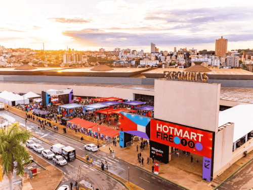 Foto aérea do centro de convenções Expominas durante o dia. O local está montado com diversas tendas coloridas e um pórtico de entrada vermelho com a inscrição "Hotmart Fire 10 anos". Há movimentação de pessoas e carros estacionados ao redor do grande pavilhão.
