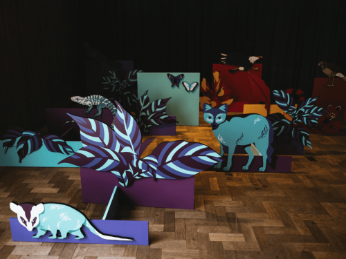 Cenografia em um ambiente interno com figuras de animais da fauna brasileira (como raposa e gambá) e folhagens coloridas espalhadas pelo chão de madeira.