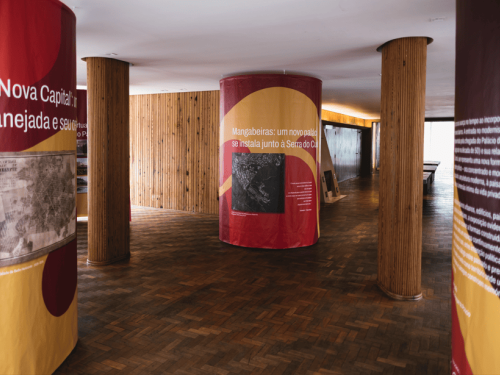 Ambiente interno de exposição cultural, com piso de madeira escura e colunas cilíndricas revestidas de madeira. Algumas colunas são envolvidas por painéis expositivos em tons de vermelho e amarelo, com textos e imagens sobre a história urbana. O espaço é amplo, bem iluminado e organizado para circulação de visitantes.