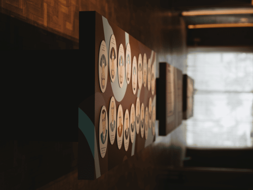 Painel expositivo em ambiente interno, disposto ao longo de um corredor com piso e paredes em tons escuros. O painel apresenta diversos círculos com textos e ilustrações, organizados de forma sequencial. Ao fundo, há uma grande janela que permite a entrada de luz natural, iluminando parcialmente o espaço.