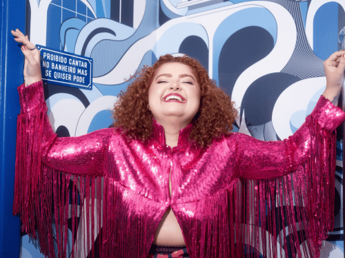 Mulher branca de cabelos cacheados e ruivos sorri com a cabeça para trás, de braços abertos, vestindo uma jaqueta rosa de paetês com franjas. Ao fundo, uma parede com estampa abstrata azul e branca exibe uma placa com os dizeres: "Proibido cantar no banheiro mas se quiser pode".