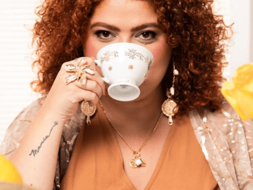 Mulher branca de cabelos cacheados e ruivos olha fixamente para a câmera enquanto bebe de uma xícara de porcelana branca com estrelas douradas. Ela veste uma blusa laranja com sobreposição bege brilhante e usa grandes acessórios dourados, como um anel de flor e brincos longos. No antebraço, exibe uma tatuagem escrita.