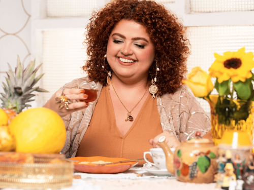 A mesma mulher sorri olhando para baixo, segurando um pequeno copo de vidro (tipo americano). Ela está sentada à mesa de café decorada com frutas amarelas (melão e abacaxi), um bule artesanal pintado e um vaso com flores amarelas.