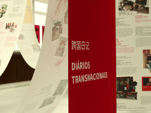 Close em um totem vermelho com o título "Diários Transnacionais" escrito em branco (em português e chinês). Ao fundo, as faixas de papel da exposição estão desfocadas.