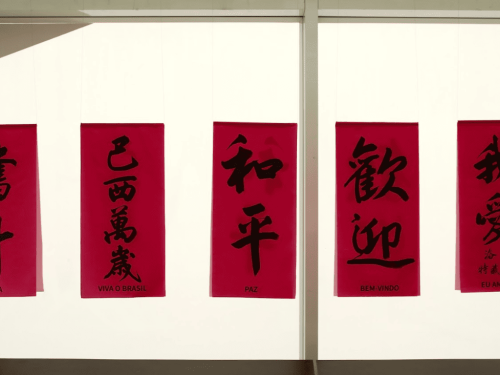 Cinco pequenos estandartes vermelhos pendurados lado a lado. Cada um traz caligrafia chinesa em preto com a tradução em português abaixo: "Luta", "Viva o Brasil", "Paz", "Bem-vindo" e "Eu amo BH".