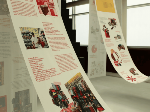 Interior de uma galeria exibindo longas faixas brancas de papel que pendem do teto em curvas. As faixas contêm textos em português e chinês, acompanhados por fotografias e ilustrações relacionadas à imigração e ao comércio.