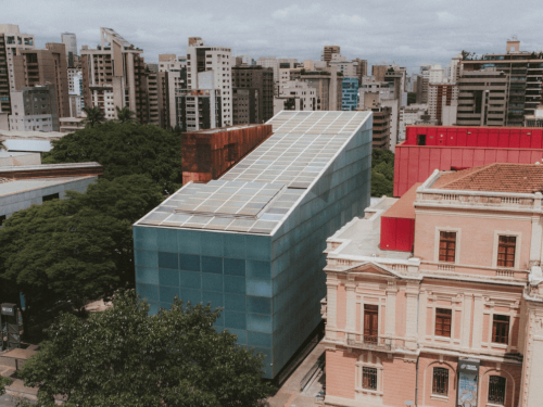 Fotografia aérea de um prédio moderno de formato inclinado com fachada azul e teto coberto por painéis solares. Ele está situado entre edifícios históricos de arquitetura clássica e uma área arborizada, com o horizonte da cidade ao fundo.