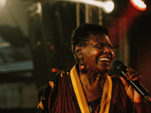 Foto colorida de Dona Eliza cantando em um palco iluminado. Ela é uma mulher negra, sorri alegremente e segura um microfone. Veste uma túnica estampada em tons de marrom e amarelo, com luzes de palco ao fundo criando uma atmosfera de show.