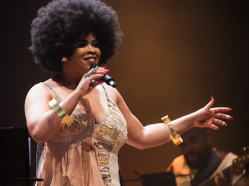 Foto de Elisa de Sena no palco. Ela é uma mulher negra com cabelo afro volumoso, veste um figurino bege com detalhes dourados e pulseiras largas. Ela segura um microfone e gesticula com a mão esquerda durante sua performance.