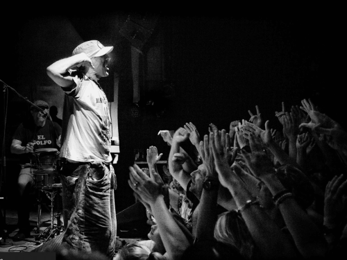 Fotografia em preto e branco de um show de Manu Chao. O cantor está de perfil no palco, usando boné e camiseta, com uma expressão vibrante enquanto interage com o público. À sua frente, uma multidão com as mãos levantadas e aplaudindo, criando uma atmosfera de energia e proximidade. Ao fundo, à esquerda, vê-se um músico tocando percussão.
