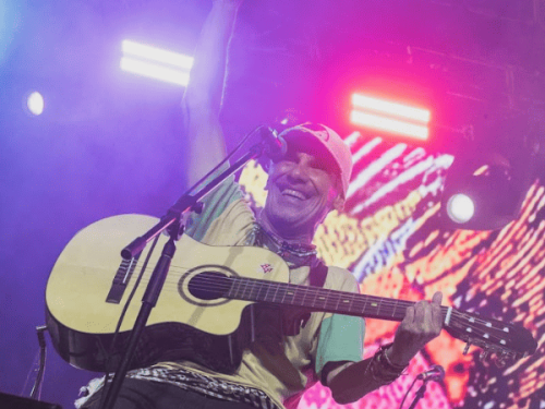 Plano médio de Manu Chao tocando um violão acústico e sorrindo, com um dos braços erguidos para o público. A cena é iluminada por luzes rosas e roxas, com fumaça de palco e um telão colorido ao fundo.