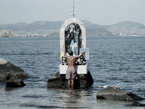 Homem preto adorando altar com escultura de santo à beira mar. Filme "Fé e Fúria".