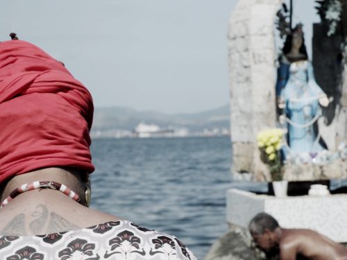Duas pessoas pretas na beira do mar. Altar com escultura de santo ao fundo com vasos de flores e imagens de santo em papel. Filme "Fé e Fúria".