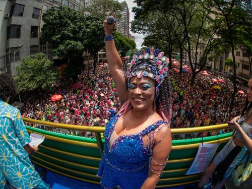 Três pessoas com fantasia de carnaval em cima do trio elétrico. Multidão ao fundo. Ensaio de Carnaval do Bloco Quando Come se Lambuza 2026