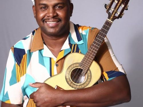 Homem negro, sorridente, está sentado de frente para a câmera, segurando um cavaquinho junto ao peito. Ele usa boina branca e camisa estampada em tons de azul, amarelo e marrom. O fundo é neutro em tom cinza.