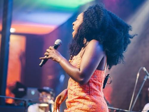 Mulher negra, de cabelos crespos volumosos, está em pé no palco cantando enquanto segura um microfone. Ela veste um vestido curto laranja com estampa discreta. O ambiente tem iluminação colorida em tons de azul, rosa e verde, e ao fundo há instrumentos musicais e equipamentos de som.