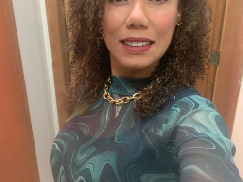 Uma mulher preta de cabelo cacheado usando maquiagem, roupa azul e colar dourado