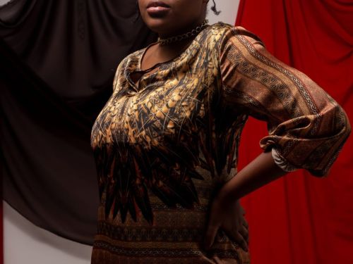 Uma mulher preta com vestido marrom e dourado escuro, e lenço na cabeça com dois panos na parede de fundo, um marrom e o outro vermelho