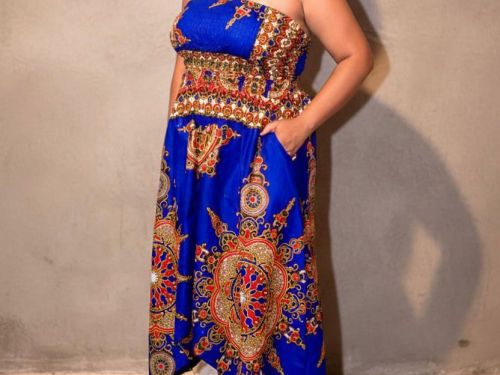 Uma mulher preta com cabelo cacheado pequeno mas volumoso, com acessórios (brinco e colar), vestido azul estampado e sandália branca, e fundo bege. 