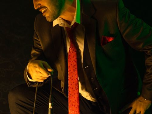 Artista Dante Alessandro cantando. Ele veste terno preto com gravata vermelha e se apoia em uma perna enquanto segura o microfone.