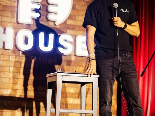 O comediante Afonso Padilha está em pé no centro de um palco de stand-up comedy. Ele é um homem branco, de barba e cabelos escuros, vestindo uma camiseta preta, calça jeans escura e tênis claros.