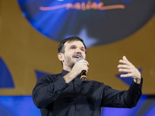 O Padre Patrick aparece em cima de um palco, vestindo uma camisa social preta e calça escura com cinto. Ele está segurando um microfone com a mão direita e gesticulando com a mão esquerda enquanto fala ou se apresenta. Ao fundo, há um painel azul e dourado com o logotipo do evento "Totus Tuus". A iluminação foca no padre, deixando o cenário levemente desfocado.