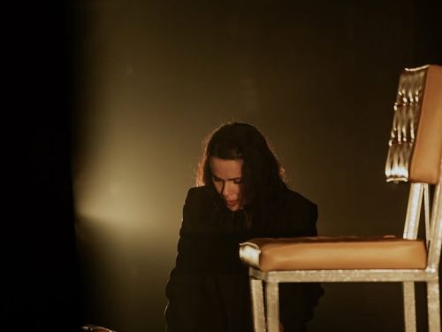 Cena teatral com a atriz Débora Falabella em um palco escuro. Ela veste roupas pretas e está inclinada sobre uma mesa, manuseando papéis ou um roteiro. À sua frente, destaca-se uma cadeira de metal com estofado caramelo. A iluminação é focada e dramática, vindo da lateral e criando um forte contraste de luz e sombra (efeito chiaroscuro), o que reforça o tom introspectivo da cena.