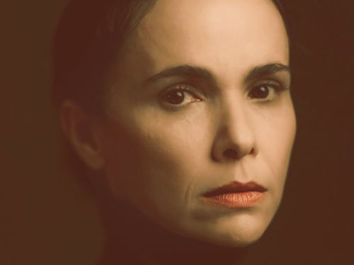 Retrato em close-up da atriz Débora Falabella. Ela aparece com uma expressão facial séria e profunda, olhando diretamente para a câmera. A iluminação é dramática, com tons quentes e terrosos (marrom e âmbar), destacando os traços de seu rosto sobre um fundo escuro e desfocado.