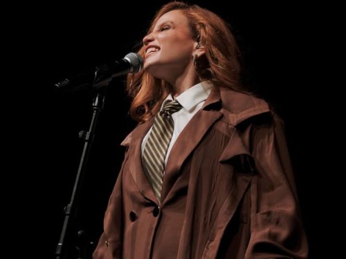 A foto é um plano médio de uma cantora ruiva, capturada de perfil lateral. Ela está sorrindo com os olhos fechados, demonstrando emoção enquanto canta em um microfone de pedestal. Ela veste um figurino clássico e elegante: um sobretudo marrom de corte largo, camisa branca e uma gravata listrada em tons de bege e marrom. O fundo é completamente preto, o que destaca a artista e os detalhes de sua expressão e vestimenta.