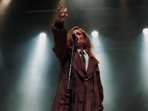 A foto mostra uma cantora de cabelos ruivos no centro do palco, vista de um ângulo de baixo para cima. Ela veste um terno e sobretudo marrom, com camisa branca e gravata. Ela está cantando em um microfone de pedestal, com o braço direito estendido para cima em um gesto expressivo. O fundo é escuro, preenchido por fumaça de palco e luzes de refletores brancos que criam uma atmosfera dramática e cinematográfica.