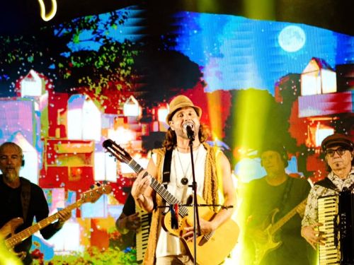 Foto colorida do grupo Falamansa em um show. No centro, o vocalista Tato sorri ao microfone com seu violão e chapéu característico. Ele é ladeado pelos músicos com baixo, guitarra e sanfona. Ao fundo, um telão exibe casinhas coloridas sob um grande letreiro de neon amarelo com o nome da banda. Feixes de luz vibrantes iluminam o palco acima da silhueta da plateia.