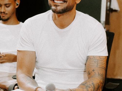 Fotografia em plano médio do cantor Diogo Nogueira sorrindo. Ele usa óculos de grau, camiseta branca e possui diversas tatuagens nos braços, incluindo uma partitura musical. Ele está sentado, segurando um microfone com as duas mãos.