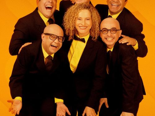 Foto promocional do grupo de comédia Os Melhores do Mundo. Os seis integrantes (uma mulher ao centro e cinco homens ao redor) posam sorridentes e abraçados contra um fundo amarelo vibrante. Todos vestem ternos pretos com camisas amarelas e gravatas escuras (a mulher usa uma gravata borboleta). A imagem transmite uma energia alegre e de cumplicidade, típica de um grupo teatral de humor.