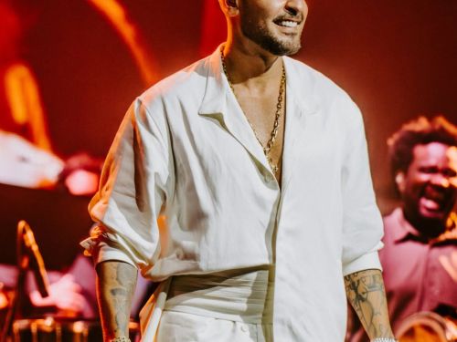 Fotografia de Diogo Nogueira em uma performance ao vivo sob luzes quentes e avermelhadas. Ele veste um conjunto de camisa e calça brancas de tecido leve e sorri enquanto segura o microfone. Ao fundo, é possível ver parte da banda e instrumentos de percussão.