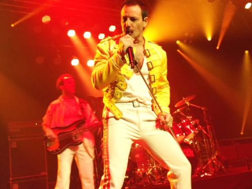 Foto de uma apresentação ao vivo do espetáculo God Save The Queen. Em primeiro plano, o vocalista Pablo Padin aparece caracterizado como Freddie Mercury, segurando o microfone e cantando com intensidade. Ele veste a icônica jaqueta amarela com fivelas e calça branca com listras laterais vermelhas. Ao fundo, é possível ver um baixista e parte da bateria sob uma forte iluminação de palco em tons de amarelo e vermelho, criando uma atmosfera vibrante de show de rock.