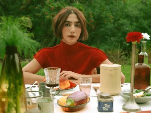 Uma mulher de cabelos curtos castanhos e blusa de gola alta vermelha está sentada ao centro de uma mesa de jantar ao ar livre. Ela olha diretamente para a câmera com uma expressão serena. A mesa, coberta por uma toalha branca, está repleta de itens diversos: copos de vidro, pratos com alimentos (incluindo uma berinjela), recipientes de cerâmica e pequenos vasos com flores. Ao fundo, há uma vegetação densa e verde, criando uma atmosfera rústica e natural.