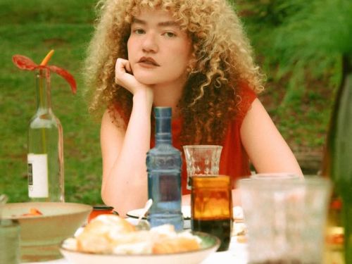 Uma mulher com cabelos loiros, volumosos e cacheados está sentada à mesa de jantar ao ar livre, apoiando o rosto em uma das mãos. Ela veste uma blusa vermelha e olha pensativa para a câmera. A mesa está posta com uma toalha branca, exibindo garrafas de vidro coloridas (azul e transparente), pratos com pães e copos. O fundo é composto por um gramado verde e folhagens, mantendo a estética rústica e natural da foto anterior.