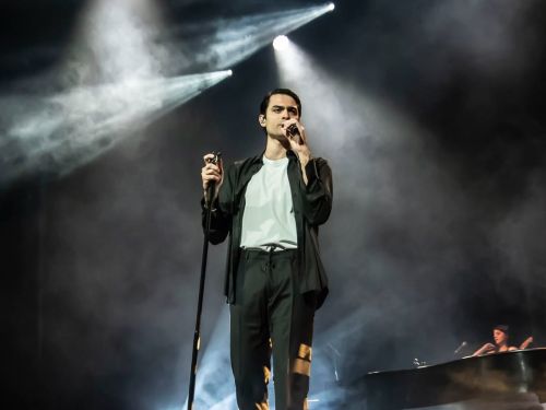Fotografia colorida do cantor Matteo Bocelli em uma performance ao vivo no palco. Ele está em pé no centro, segurando o microfone com a mão direita e cantando. Veste calça preta, camiseta branca e uma camisa preta aberta por cima. O palco está envolto em fumaça cenográfica e iluminado por fortes feixes de luz branca que descem do topo. À direita, ao fundo, é possível ver parte de um piano de cauda e uma musicista.