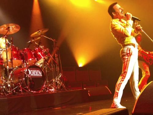 Foto de perfil do palco durante o show God Save The Queen. À direita, o vocalista está inclinado para trás, segurando o pedestal do microfone em uma pose triunfante. Ele veste a famosa jaqueta amarela e calça branca com listras vermelhas. À esquerda, em destaque no fundo, o baterista toca um kit de bateria vermelho que exibe o logotipo do espetáculo no bumbo. A cena é banhada por uma iluminação dourada intensa, capturando a energia de uma performance de rock clássico.